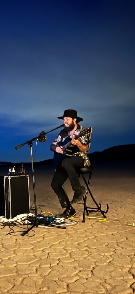 David Rice performing live outdoors at dusk in the desert with guitar and microphone